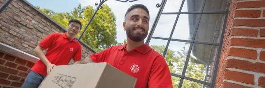 Técnicos de MediaMarkt recogiendo una caja en un domicilio, vistiendo camisetas rojas y sonriendo.