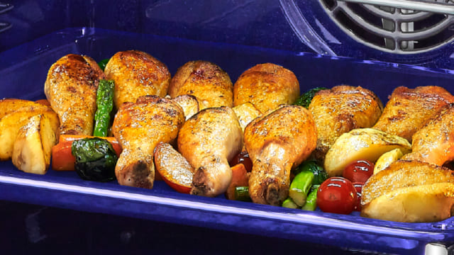 En un horno azul, hay pollo asado con patatas, tomates y verduras verdes.