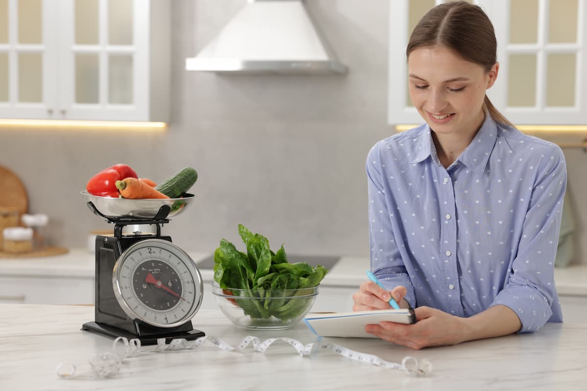 Mujer sonriente anotando en una tableta junto a una báscula de cocina con verduras en una cocina blanca.