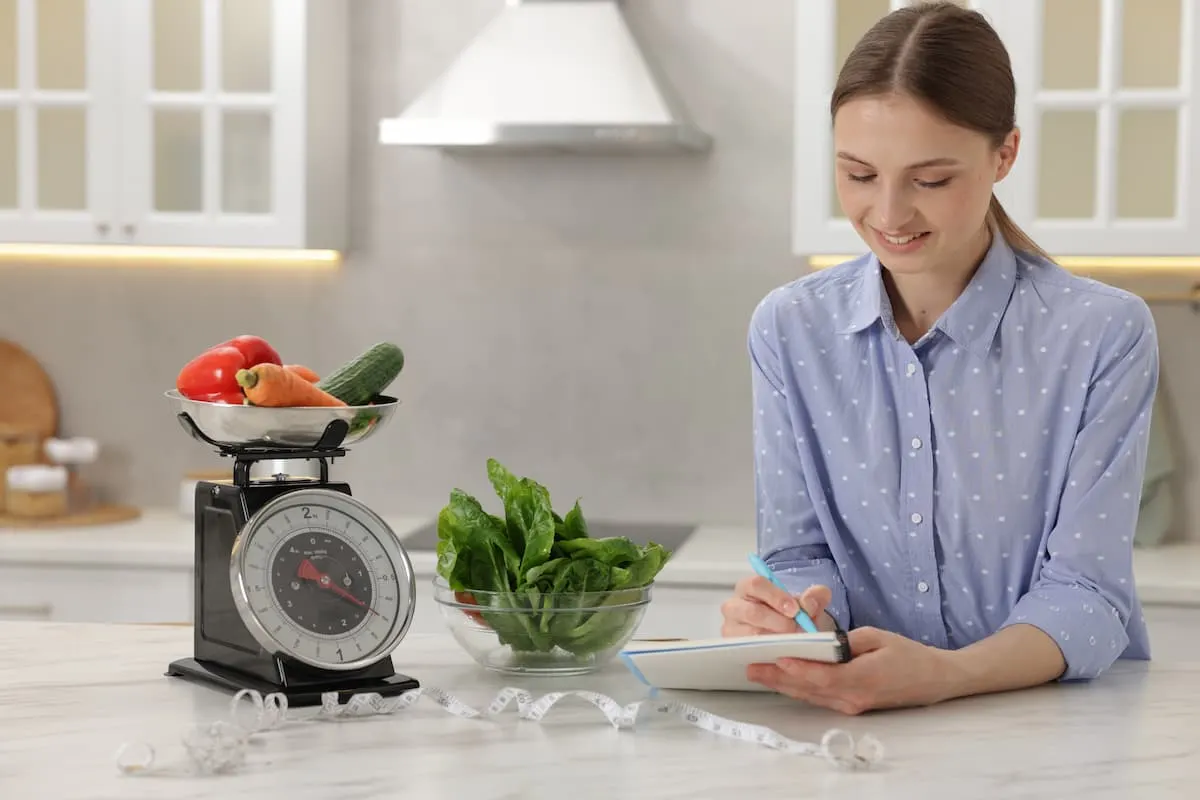 Mujer sonriente anotando en una tableta junto a una báscula de cocina con verduras en una cocina blanca.