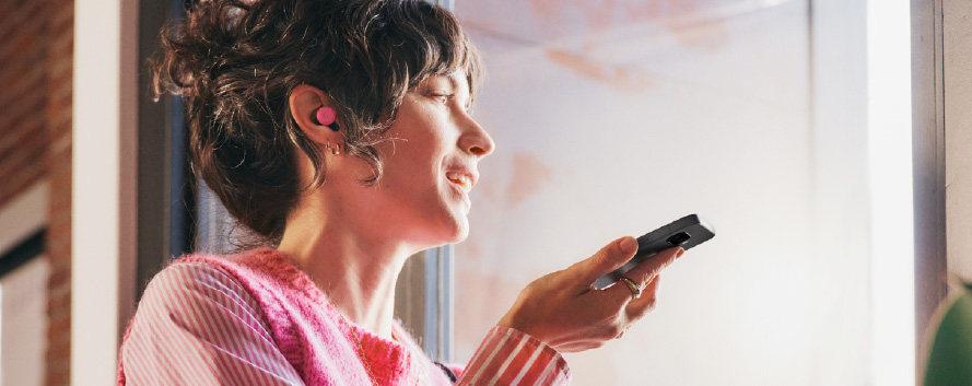 Un hombre con camisa naranja sostiene un móvil blanco junto a un vaso dorado, un cuaderno y un portátil sobre una mesa.
