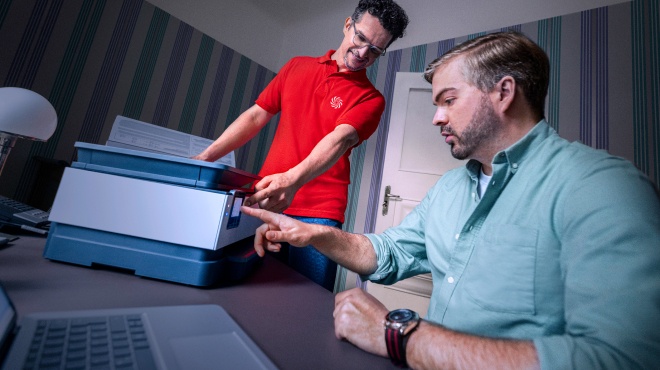 Un hombre con camisa verde habla con una mujer de camisa roja que sostiene una tableta en una habitación con plantas y lámparas.