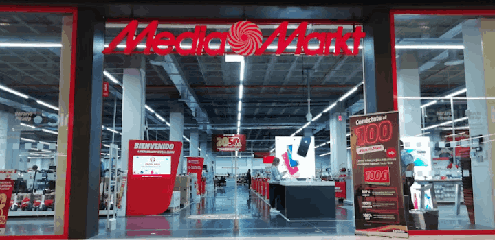 Entrada a tienda Media Markt con letrero rojo y blanco. Interior con pasillos largos, estantes con productos y carteles promocionales.