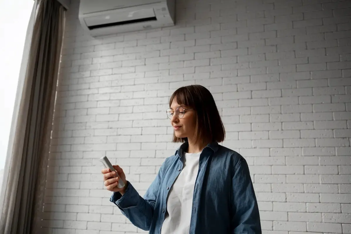 Mujer con gafas y camisa azul sosteniendo un control remoto frente a una pared de ladrillo blanco con aire acondicionado y cortinas.