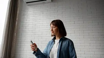 Mujer con gafas y camisa azul sosteniendo un control remoto frente a una pared de ladrillo blanco con aire acondicionado y cortinas.