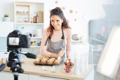 Una mujer con delantal prepara magdalenas y fresas frente a una cámara en una cocina luminosa.