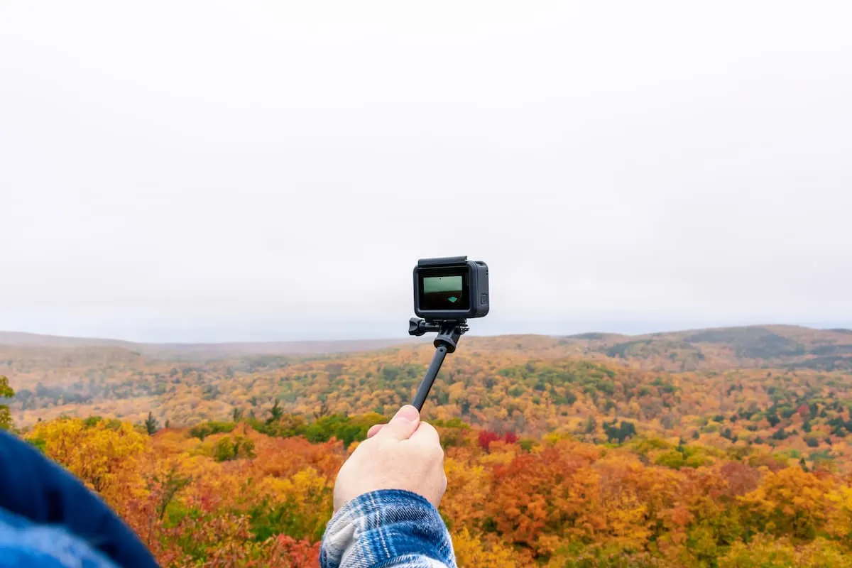 Mano sosteniendo una cámara GoPro en palo selfie frente a un bosque otoñal.