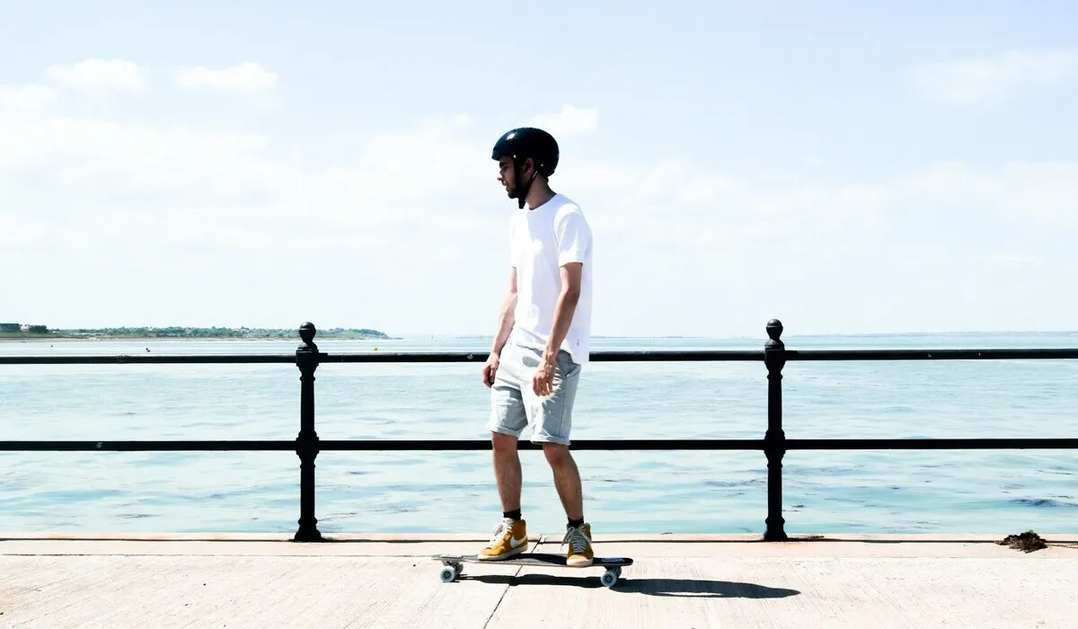 Un hombre con casco negro y ropa clara está en una patineta junto a una barandilla negra, con el mar y el cielo nublado de fondo.
