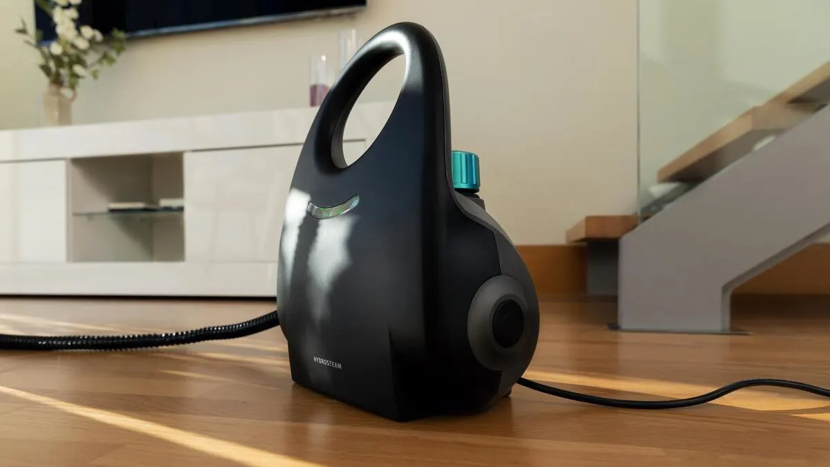 Aparato de limpieza a vapor negro con asa y tapa azul, sobre suelo de madera. En segundo plano, mueble y escalera blancos.
