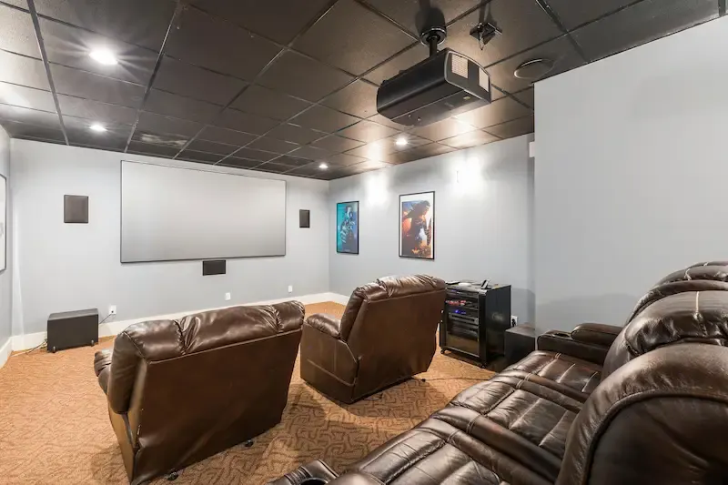 Sala de cine con pantalla grande, butacas de cuero marrón y proyector en el techo oscuro. Hay altavoces y equipo de sonido.