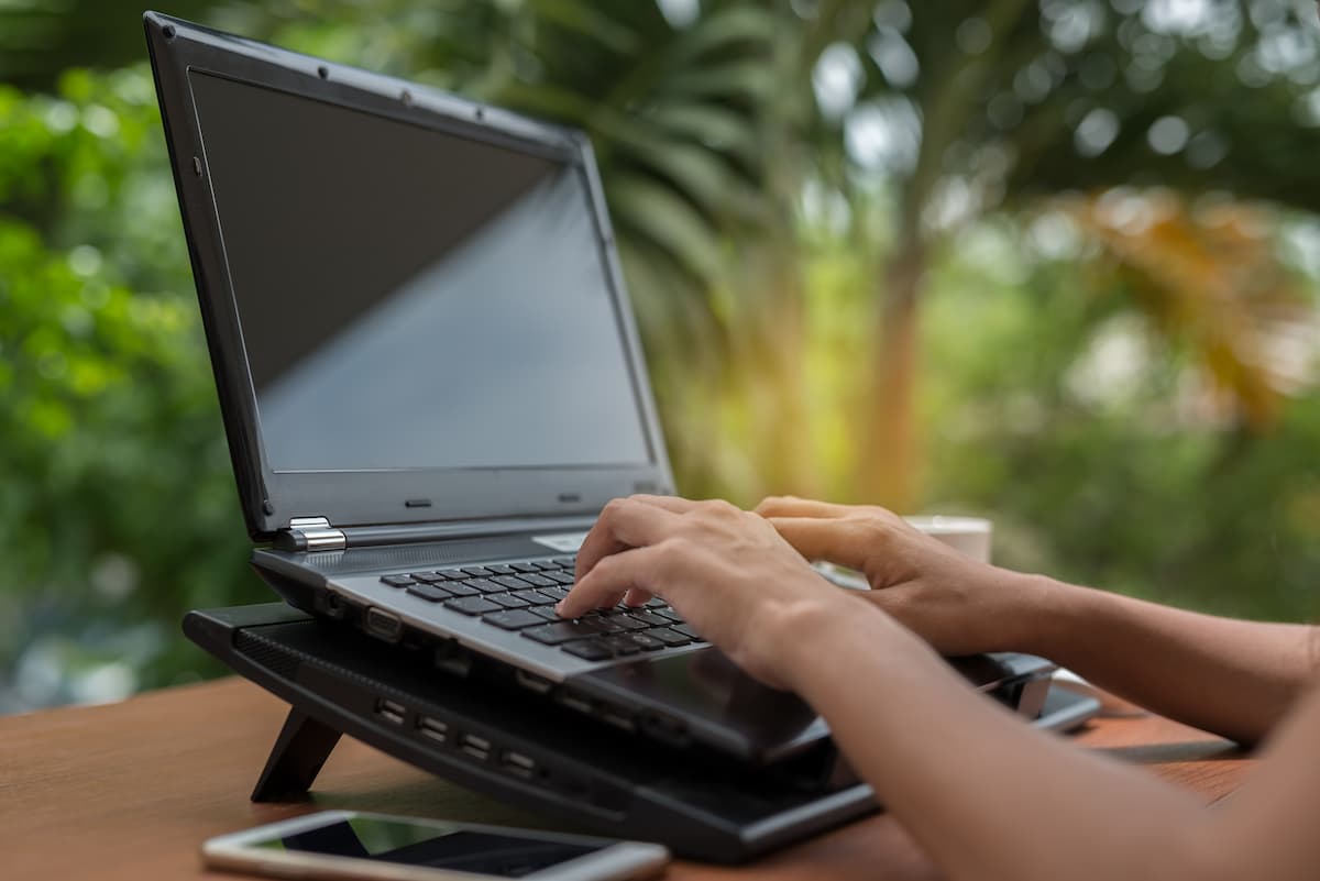 Manos tecleando en un portátil negro colocado sobre una base elevadora en una mesa de madera, con fondo desenfocado de vegetación al aire libre.