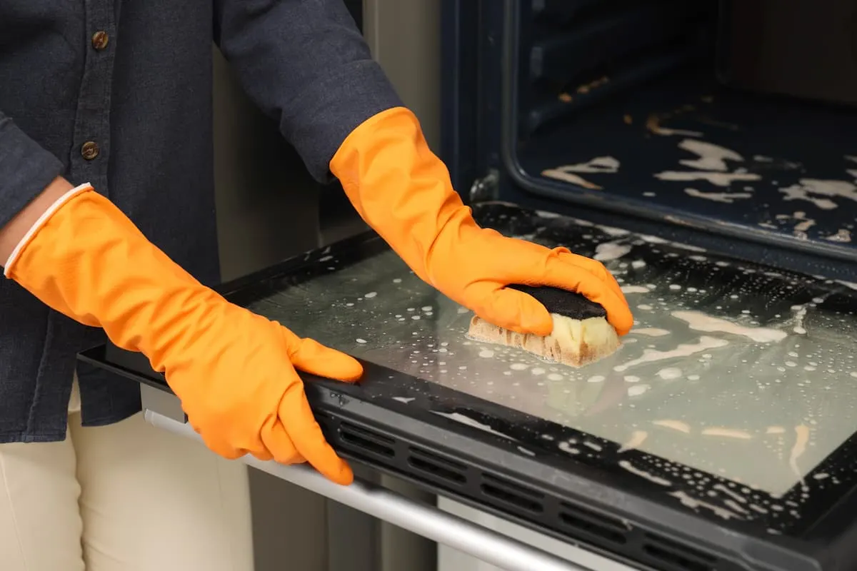 Persona con guantes de goma naranjas limpiando con una esponja la puerta de vidrio de un horno cubierta de espuma.