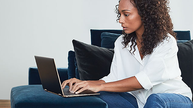 Una persona sentada en un sofá azul usa una computadora portátil. Hay cojines negros detrás. La pared es blanca.