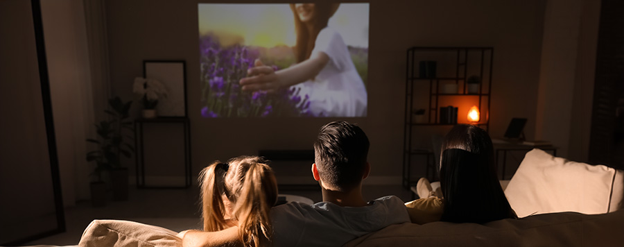 Una niña y un hombre ven una proyección en la pared en un salón oscuro. Se ve una estantería y una lámpara encendida.