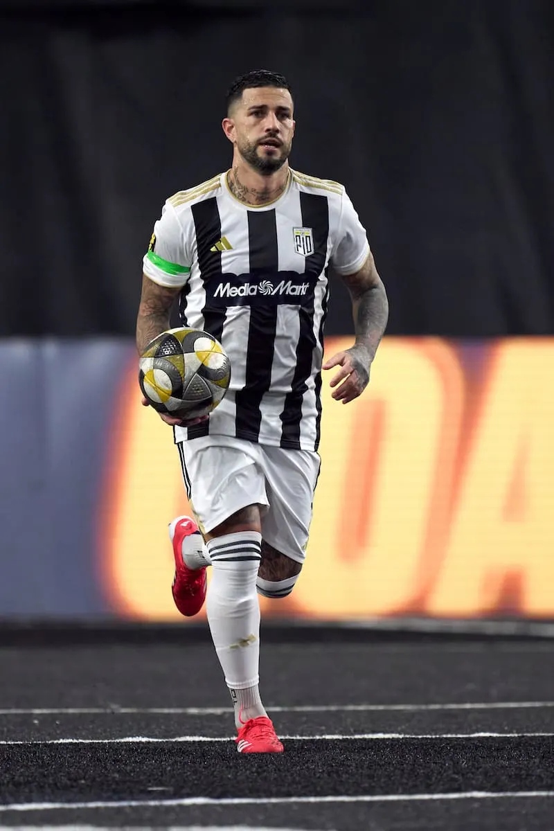 Jugador de fútbol con uniforme blanco y negro, sosteniendo un balón multicolor, corriendo sobre césped oscuro. Fondo borroso.