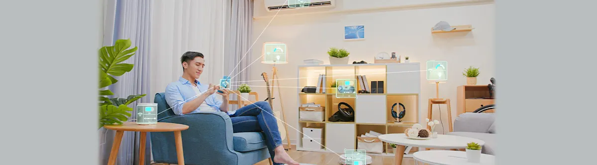 Un hombre usa un móvil en un sillón azul, con objetos conectados a través de interfaces digitales en una sala de estar luminosa.