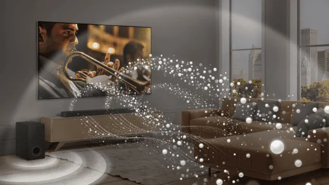 Salón con TV mostrando un trompetista, barra de sonido y altavoz. Un sofá marrón con cojines grises y ventanas al fondo.