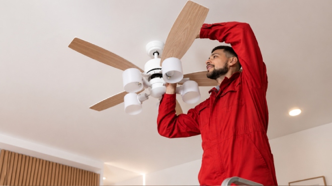 Técnico con uniforme rojo instalando un ventilador de techo blanco con aspas de madera en una vivienda moderna.