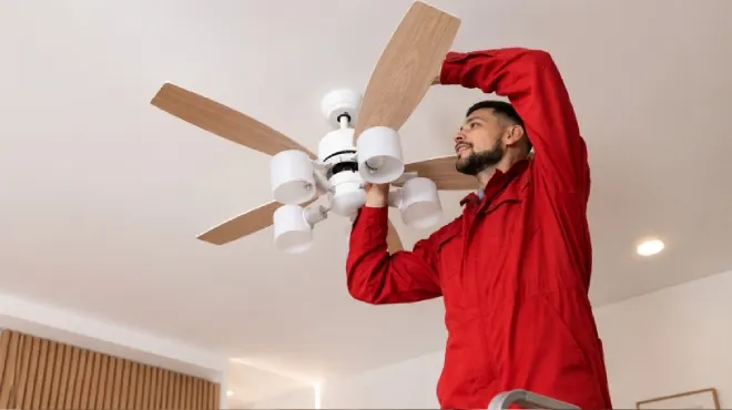 Técnico con uniforme rojo instalando un ventilador de techo blanco con aspas de madera en una vivienda moderna.