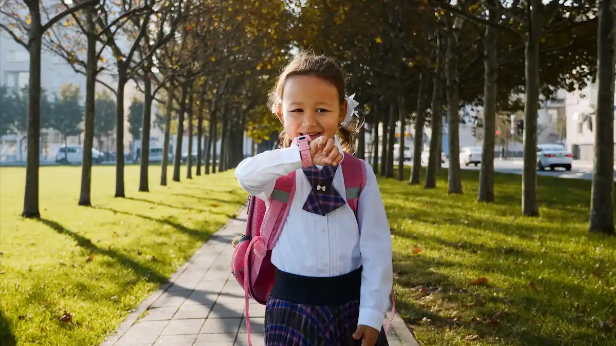 Una niña con uniforme escolar y mochila camina por un sendero arbolado, mirando un reloj rosa en su muñeca.