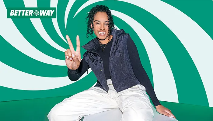Mujer sonriente con chaleco acolchado oscuro y pantalones blancos, sentada frente a un fondo verde y blanco con formas curvas, mientras hace el gesto de la paz con una mano. En la esquina superior izquierda aparece el logotipo «BETTER WAY».
