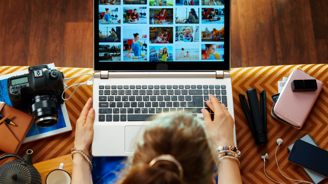 Persona usa laptop con fotos. Cámara, libreta, móvil y taza están cerca en la mesa.
