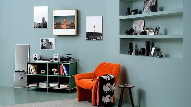 Cuarto con pared azul, estantes empotrados, sillón naranja con manta, taburete y estantería con libros y fotos enmarcadas.