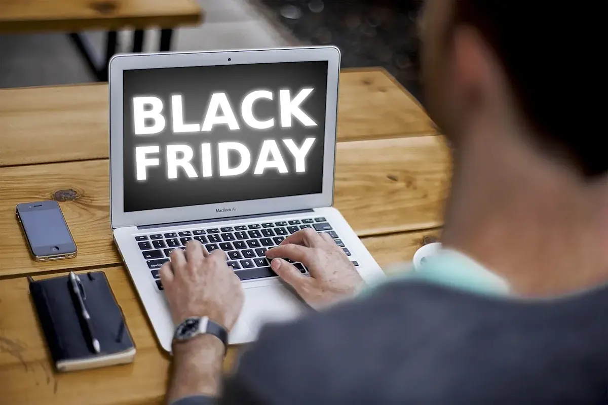 Persona usa un portátil con el texto "Black Friday" en la pantalla, junto a un móvil, una libreta y un bolígrafo sobre una mesa de madera.