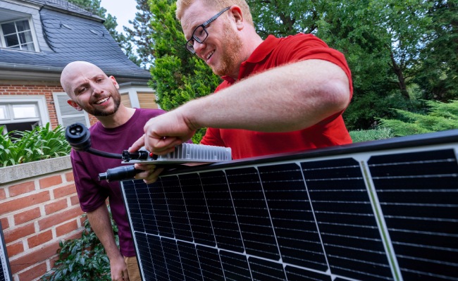 Dos hombres instalan un panel solar. Uno conecta cables, el otro mira. Fondo con ladrillos, casa y árboles.