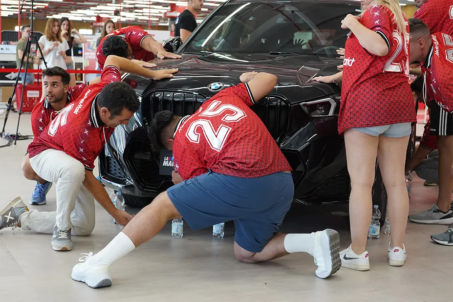 Un grupo de personas en camisetas rojas examinan un coche negro en un espacio interior bien iluminado.