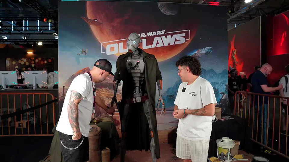 Dos hombres cerca de un robot de Star Wars, frente a un cartel promocional del juego "Outlaws", en un evento.