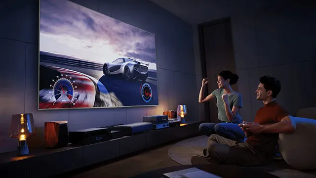 En salón, dos personas ven carrera de coches en pantalla grande. Hay lámparas y equipos electrónicos en un mueble bajo.