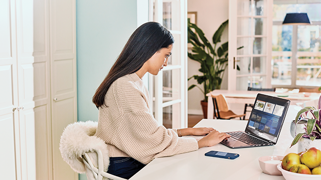 Mujer en un escritorio blanco usando una laptop. Un teléfono, fruta y una planta están cerca. Un armario blanco está al fondo.