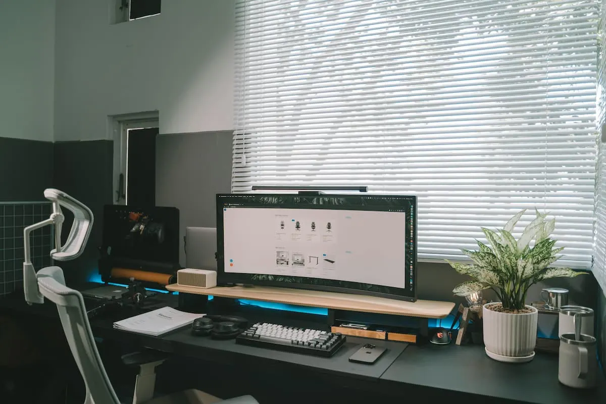Escritorio con silla ergonómica blanca, monitor curvo, teclado mecánico y planta junto a persianas cerradas.
