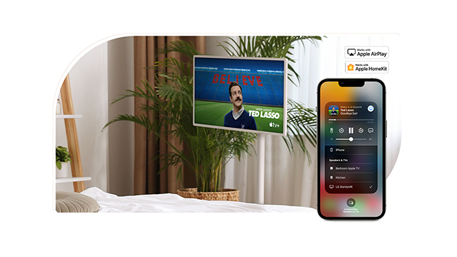 Televisión mostrando "Ted Lasso", planta grande y teléfono con panel de control, en un entorno doméstico neutro.