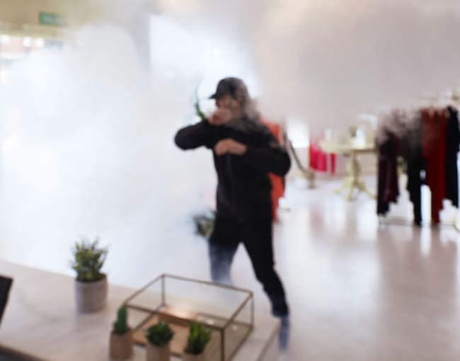 Interior de tienda con humo espeso; persona vestida de oscuro cubriéndose la cara mientras camina, junto a mesas con plantas decorativas en primer plano desenfocado.