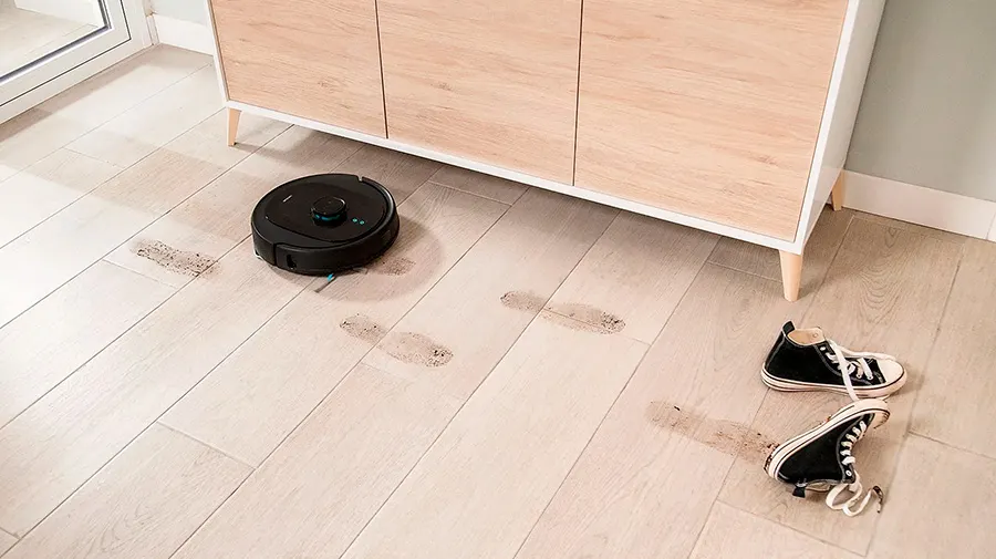 Robot aspirador negro cerca de huellas y un mueble de madera clara. Un par de zapatillas negras con cordones blancos están en el suelo.