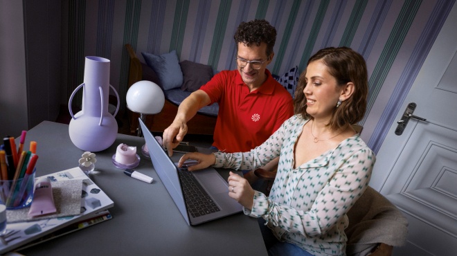 Una mujer con polo rojo sostiene un mando a distancia junto a un hombre con camisa verde frente a una televisión.