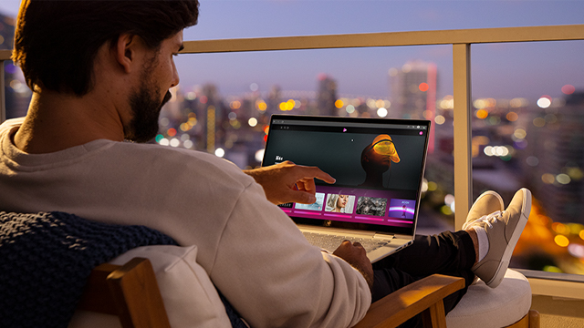 Un hombre usa una laptop en un balcón con vista a una ciudad nocturna. Él está sentado en una silla, con los pies sobre un cojín.