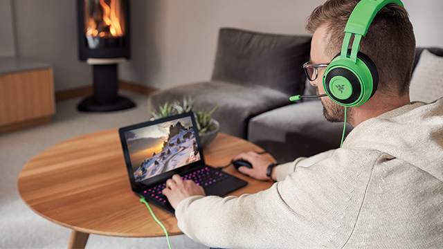 Persona con auriculares verdes usando un portátil sobre una mesa redonda de madera. En el fondo, una chimenea y un sofá gris.