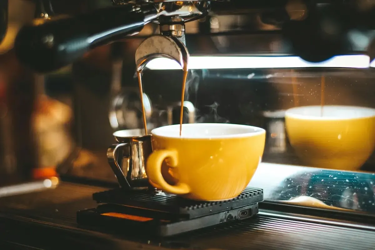 Máquina de café vertiendo líquido en taza amarilla con jarra metálica al lado, sobre base negra. Humo visible.