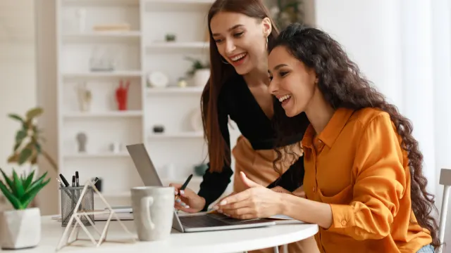 Dos mujeres sonríen mientras miran una laptop sobre una mesa blanca con una taza, un portalápices y una planta. Al fondo, estantería blanca.