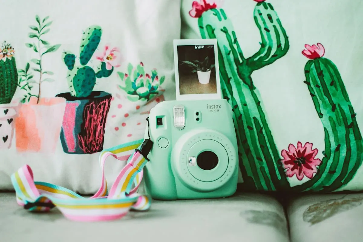 Cámara instantánea verde menta con correa de colores sobre cojines de cactus.