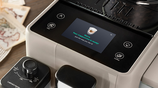 Máquina de café beige y negra con pantalla táctil que indica "Tu capuchino está listo". A la izquierda, un accesorio de leche negro.