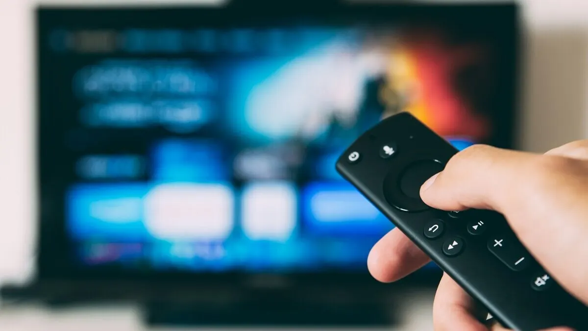 Mano sujetando un mando a distancia negro frente a un televisor con una pantalla azul brillante.