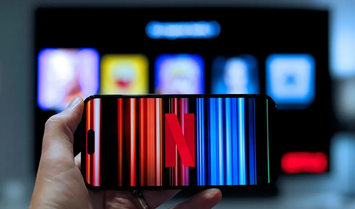 Una mano sostiene un smartphone con el logo de Netflix en la pantalla, mientras una TV desenfocada muestra aplicaciones de streaming.