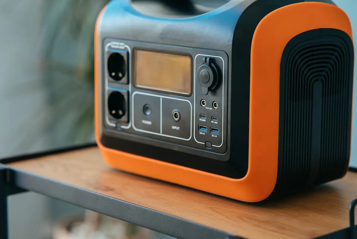 Estación de energía portátil con carcasa negra y naranja colocada sobre una mesa de madera, mostrando en primer plano sus tomas de corriente, puertos USB y panel de control frontal.