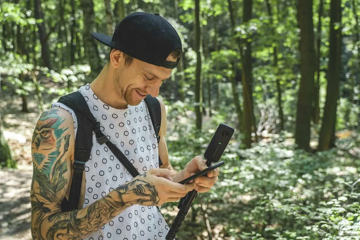 Hombre joven con cámara 360 en un bosque revisando su móvil.