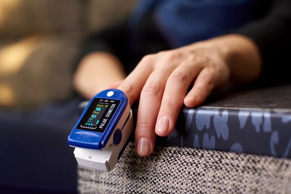  Primer plano de una mano apoyada en un sofá usando un pulsioxímetro azul en el dedo, mostrando en pantalla los niveles de oxígeno en sangre y pulso.