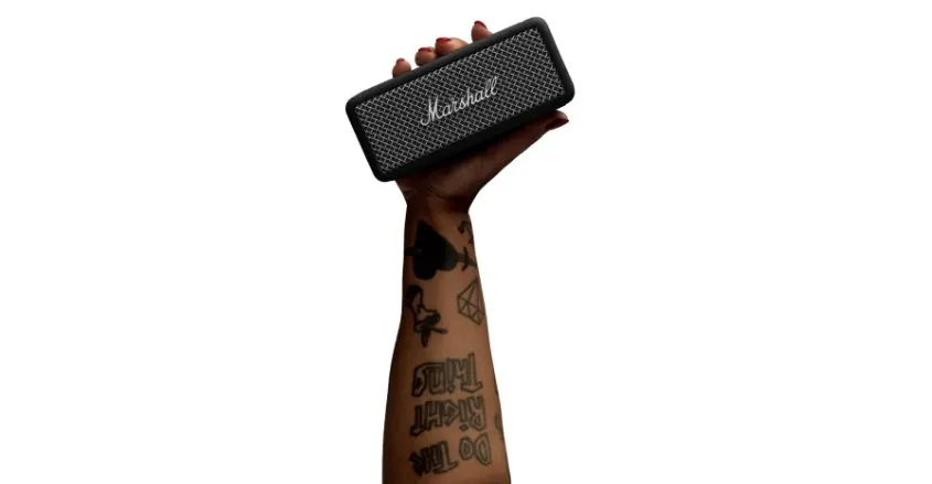 Mano con tatuajes sosteniendo un altavoz portátil Marshall negro sobre fondo blanco.